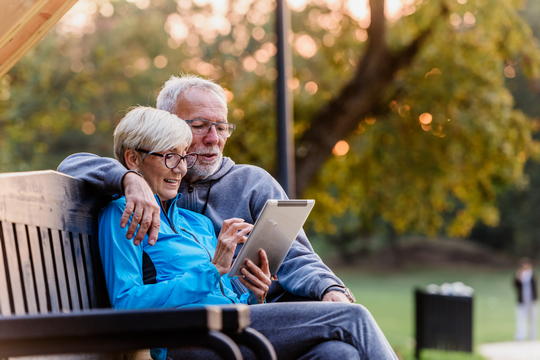 Older couple sitting cosily on a park bench looking at a digital device