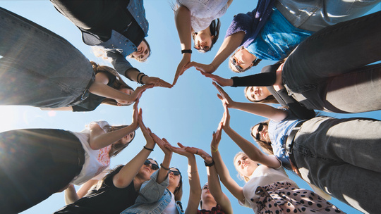 Group of people standing in a circle outdoors, creating a heart shape with their hands against a blue sky.